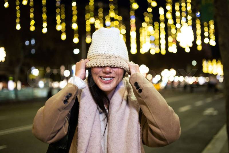 Funny woman covering her face with a warm hat in the street. Playful happy girl wearing knitted hat and scarf at night in the street.