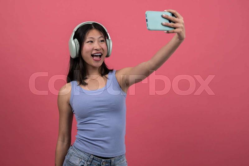 Isolated young female laughing listening to music taking a picture with smartphone. Woman with headphones and phone taking a selfie on a red background. 
