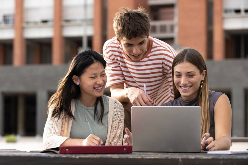 Diverse group of people working together with a laptop in campus.Young happy friends studying outside using computer and notes.