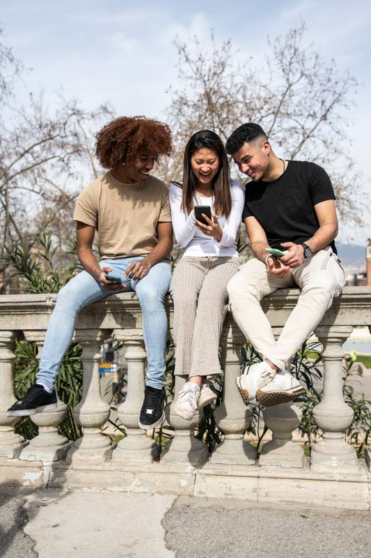 Three young diverse people looking at a smartphone sitting on a stone balcony .Two young men smiling and watching at the phone of a happy woman.