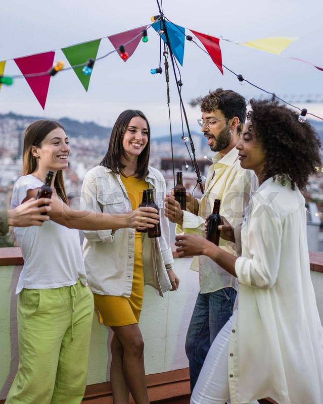 Diverse group of friends having fun and drinking beer in a rooftop party. Multiracial people toasting with bottles at evening in a terrace