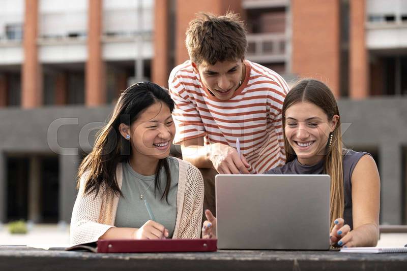 Diverse group of people working together with a laptop in campus.Young happy friends studying outside using computer and notes.