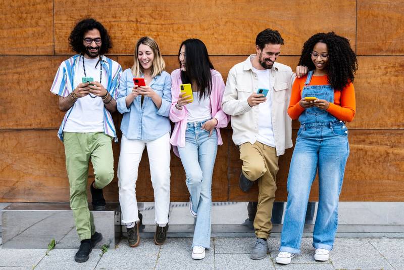 Diverse young confident people smiling and using their phones outside in a city while leaning against a wooden wall. Multiracial group of friends texting on their smartphones 