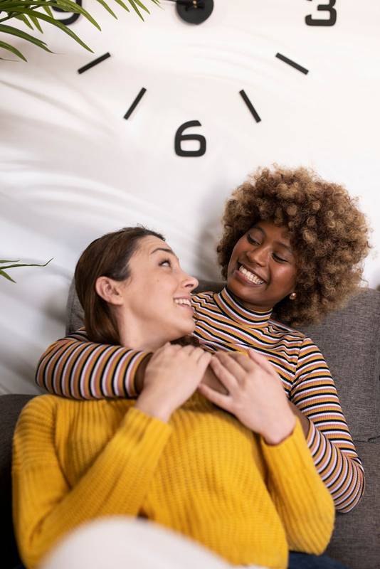 Multiracial lesbian couple hugging and laughing and relaxing in a living room. Smiling gay females couple having fun at home embracing each other on a sofa.
