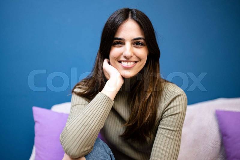 Happy young woman sitting on sofa at home and looking at camera. Portrait of comfortable caucasian woman similing and relaxing on armchair