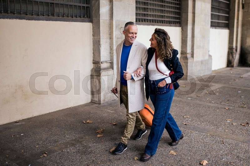 Mid adult tourist couple walking with luggage in the street. Happy senior man and woman traveling with a trolley visiting a city together.