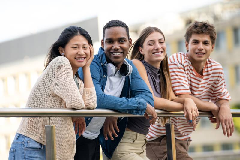 Beautiful young men and women smiling and looking at camera leaning on a balcony. Diverse group of cheerful and hopeful millennial people staring relaxed at camera.