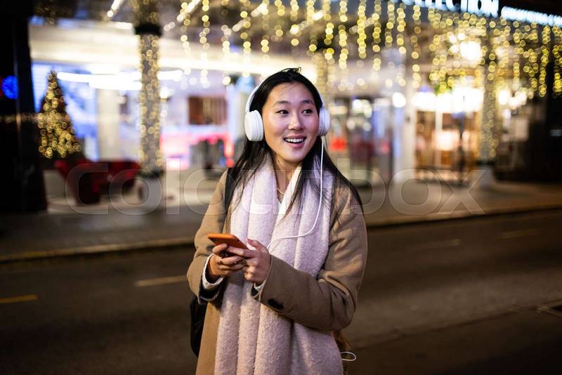 Young relaxed girl texting and wearing headphones with christmas lights behind. Happy woman listening to music at the street during winter using her phone.