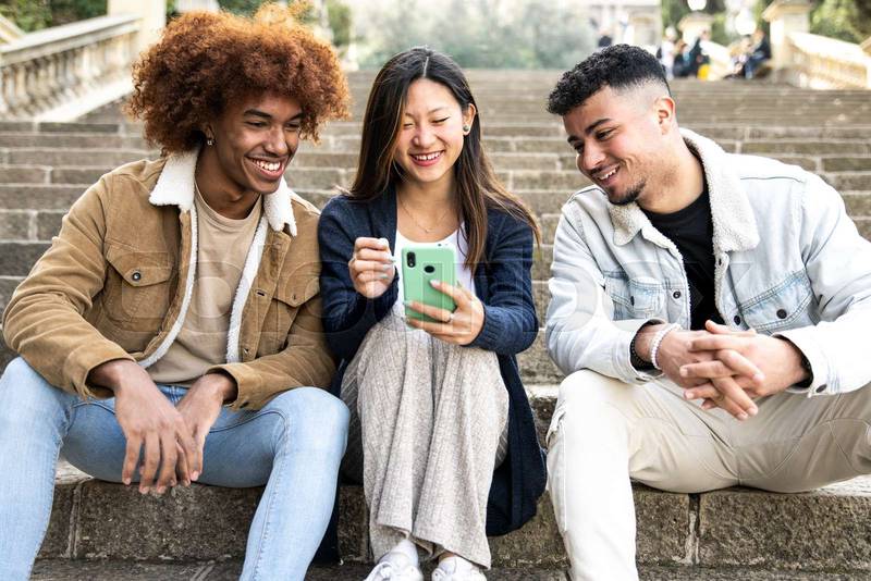 Three young diverse people looking at a smartphone sitting on the stairs.Two young men smiling and watching at the phone of a happy woman.