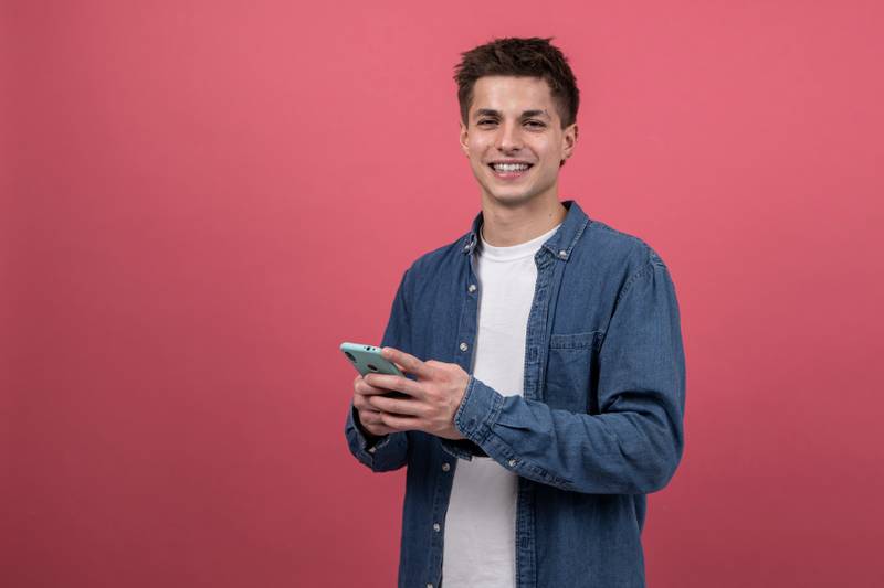 Joyful and satisfied man texting with phone looking to the camera. Smiling young male using online smartphone isolated in a red background.
