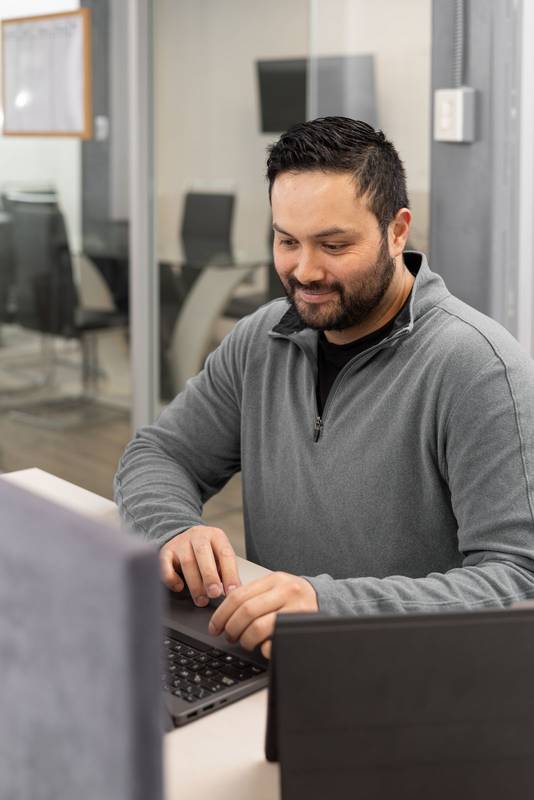 man with a beard wearing a gray blazer types on his laptop at a desk. He is concentrated and working