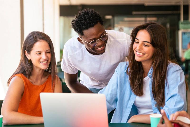 Group of happy multiracial students sitting in a cafe bar looking at laptop. Three cheerful friends studying together with a computer