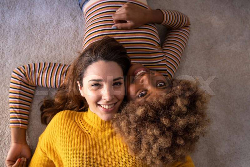 Top view of lovely multiracial lesbian couple lying on a carpet looking at camera. Beautiful happy homosexual women smiling and relaxing together on the floor.
