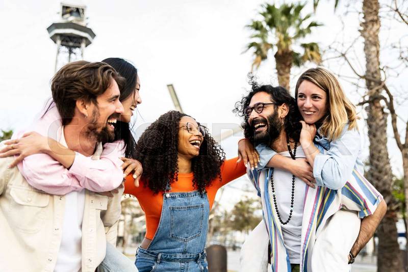 Multiracial group of friends giving piggyback ride and looking each other. Diverse young women and men having fun laughing and carefree staring at each other outside.
