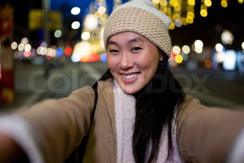 Joyful young woman taking a selfie at night with beautiful christmas lights. Beautiful cheerful female taking a picture in the street wearing a hat.