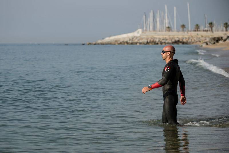 Swimmer wearing neoprene entering the water in an urban beach. Athlete ready for swimming in the ocean with wetsuit and glasses.