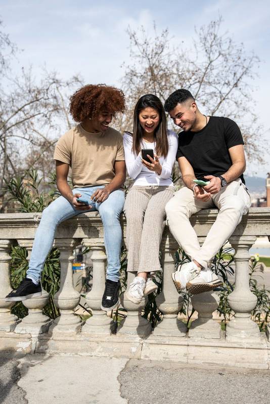 Three young diverse people looking at a smartphone sitting on a stone balcony .Two young men smiling and watching at the phone of a happy woman.