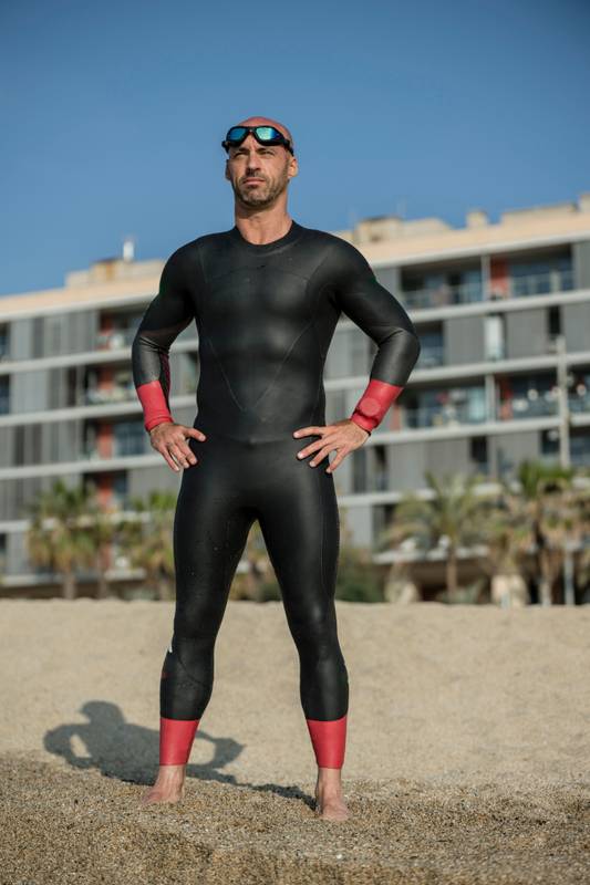 Confident man wearing a wetsuit standing in the shore ready for swimming. Professional athlete staring at the ocean in the beach with urban background.