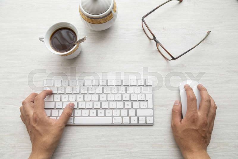 hands of a person typing on a modern keyboard and using a mouse, around them a pair of glasses, a cup of coffee
