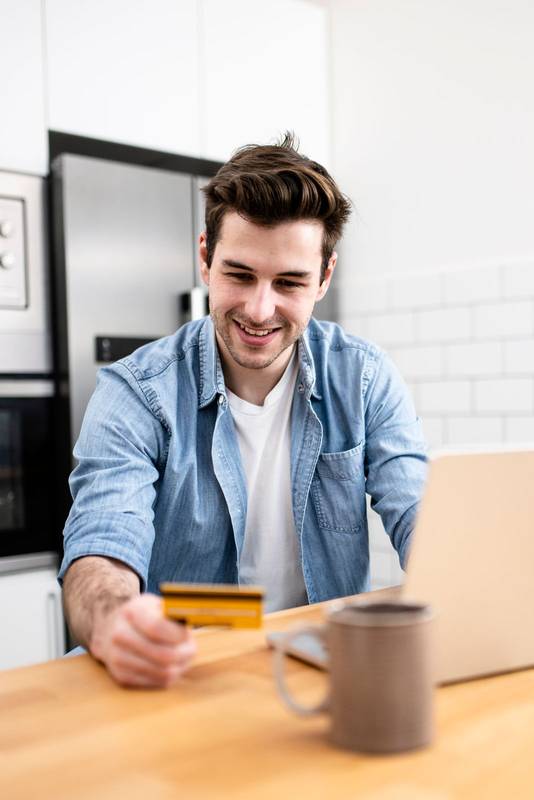 Man sitting in front of the computer holding a credit card at home - Young adult doing a online payment with his laptop at kitchen - business, technology concept