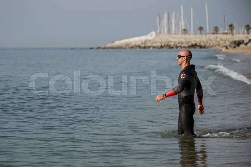 Swimmer wearing neoprene entering the water in an urban beach. Athlete ready for swimming in the ocean with wetsuit and glasses.