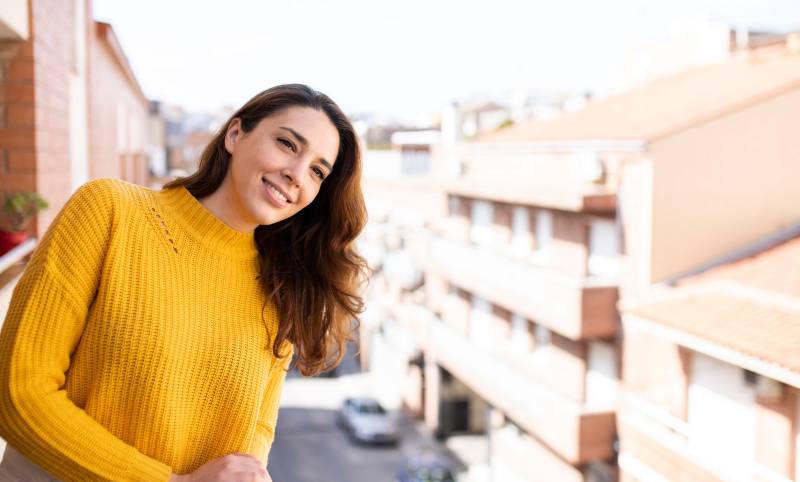 Portrait of young smiling woman looking away standing with a city background. Cheerful female looking satisfied on a balcony.