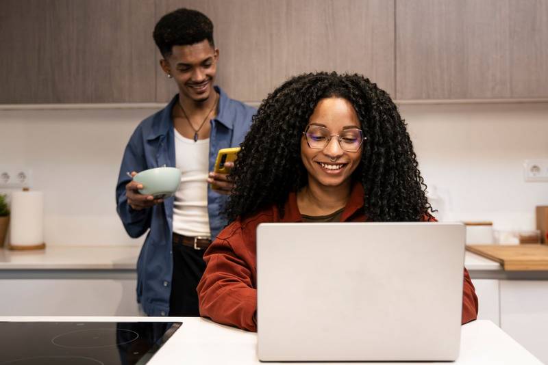 Diverse carefree couple planing together with computer and smartphone at home. Smiling man texting with phone while woman using laptop in the kitchen.
