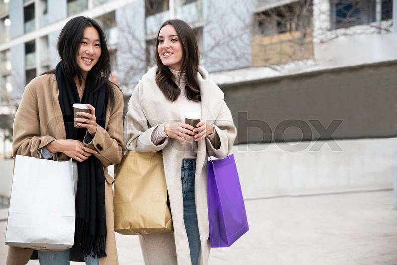 two happy young beautiful women walking on the street holding shopping bags ang cup of coffee. cheerful friends enjoying together in the mall.