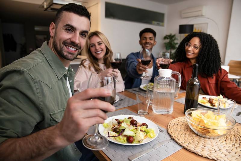 Cheerful diverse group of happy friends having party diner at home. Smiling man toasting with people eating and drinking on a modern apartment. 