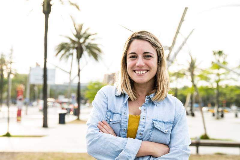 Portrait of beautiful happy young woman smiling and looking at camera with arms crossed in the street. Gorgeous cheerful blond hair female confident and satisfied standing outside in a casual style.