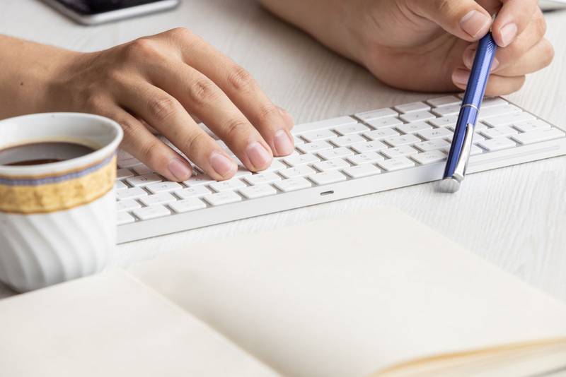 desk with a person's hands holding a pen next to a keyboard, winged a notebook with blank sheets, business in the workplace