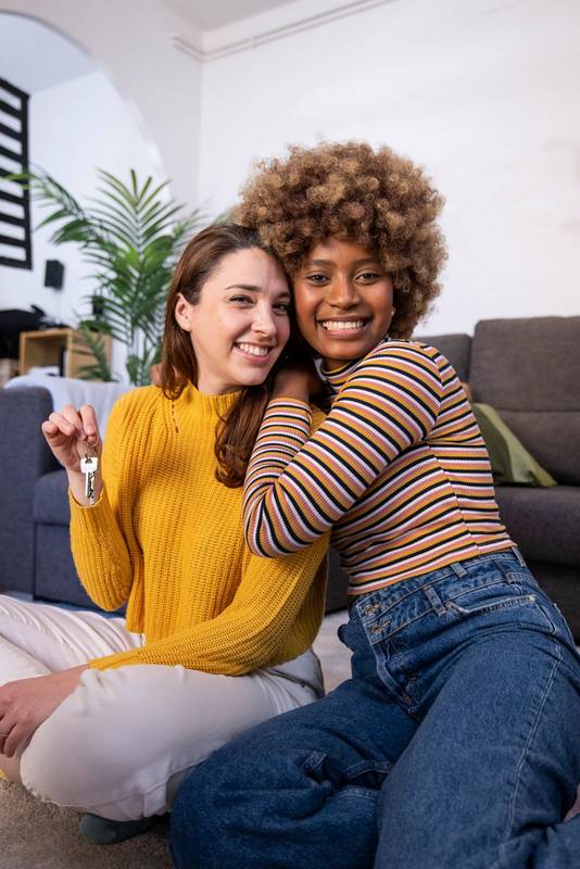 Happy multiracial lesbian couple holding keys one of them embracing her girlfriend at her new home. Two cheerful multiethnic women showing the keys of their apartment sitting on the floor.
