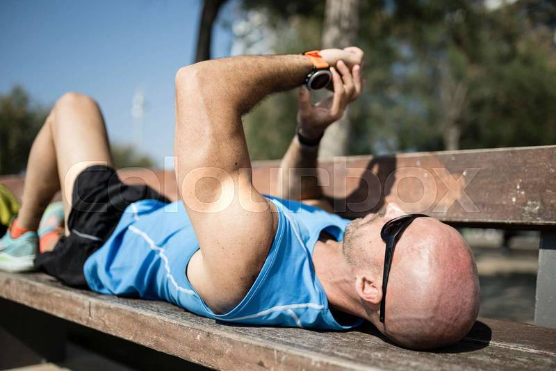 Athletic male runner training and checking his watch workout in a park. Fit man resting after training in a bench outdoors.