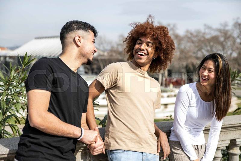 Multiracial group of young laughing people standing on a park. Three diverse happy friends having fun together outside.