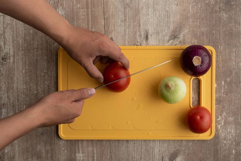 hands of a person cutting with a sharp knife a tomato in half on a colorful chopping board