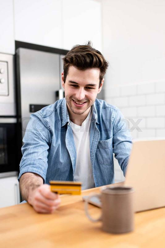 Man sitting in front of the computer holding a credit card at home - Young adult doing a online payment with his laptop at kitchen - business, technology concept