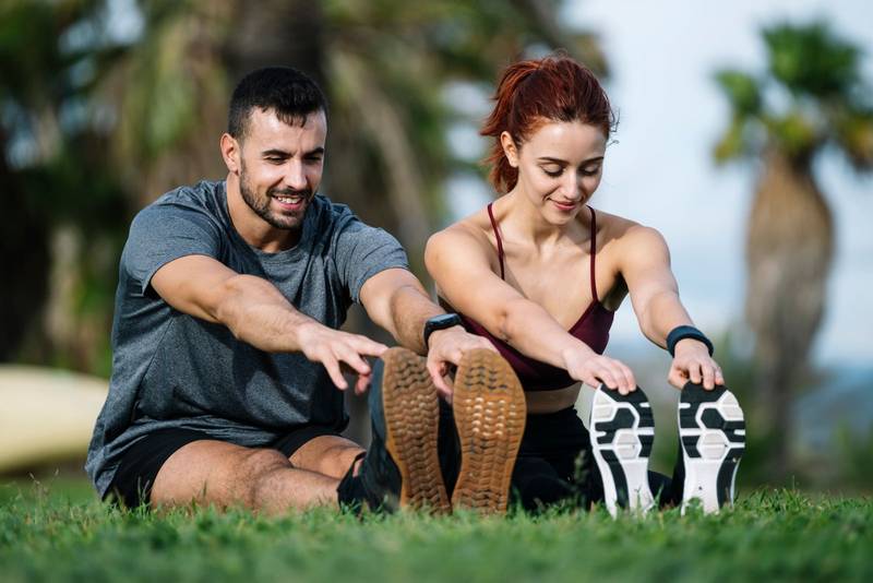 Happy young adult couple stretching in the floor after running together in the park. Smiling satisfied athletic male and female sitting in the grass in sportswear training workout outside. 
