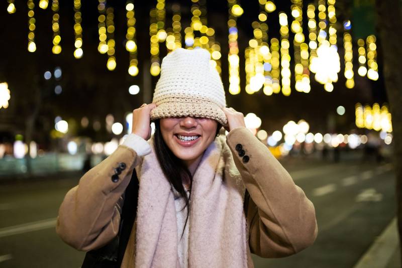 Funny woman covering her face with a warm hat in the street. Playful happy girl wearing knitted hat and scarf at night in the street.