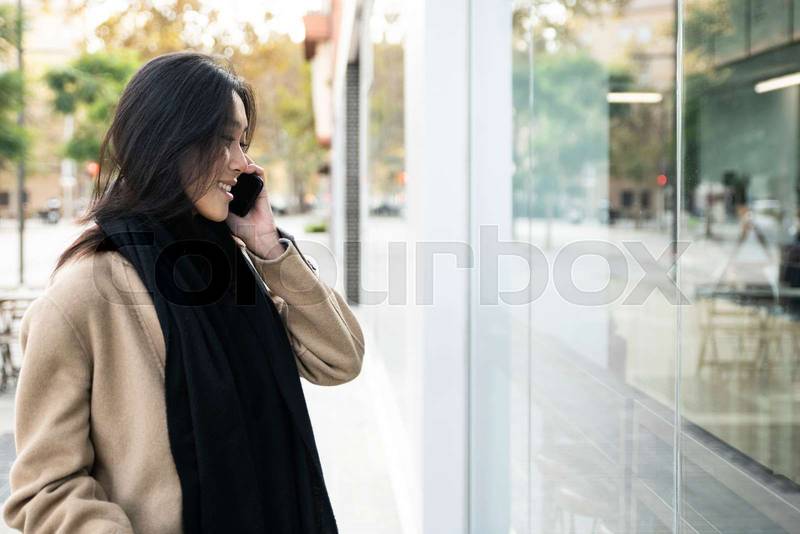 Young beautiful woman speaking smartphone and looking to showcase shop in the city. smiling shopper in front of a store