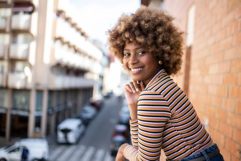 Portrait of smiling woman standing in a balcony with city background. Girl staring at camera with a smile in a high window.
