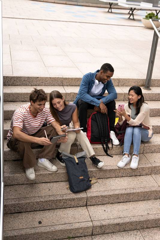 Multiracial young students studying sitting on stairs.Diverse group of happy people reading files outside.