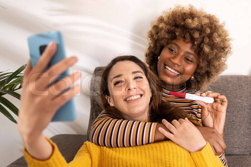 Diverse female couple taking a selfie with their positive pregnancy test in a sofa. Happy lesbian women announcing a pregnancy on their social media in a living room.