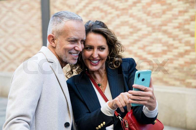 Happy mid adult tourists travelling together using phone together in the street. Lovely mature friends taking picture together while traveling.