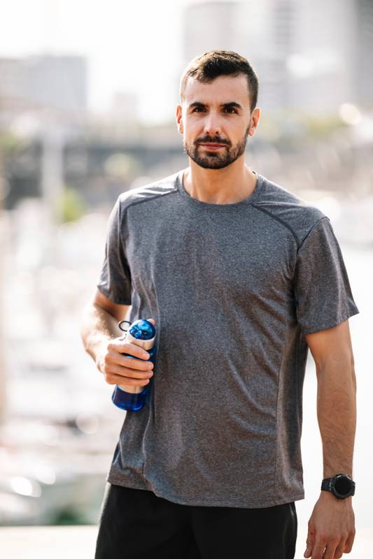 Sports fit man drinking water from bottle after training outdoor. Strong active male with recovery drink outside looking at camera.