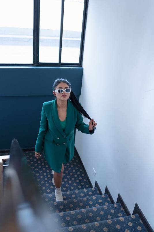 woman with ponytail wearing a jacket-style dress and sunglasses walking up some stairs indoors