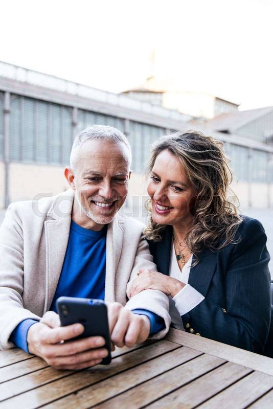 Mid adult couple looking at smartphone while sitting at outdoors table. Carefree senior man and woman using mobile phone together at a terrace in the street.