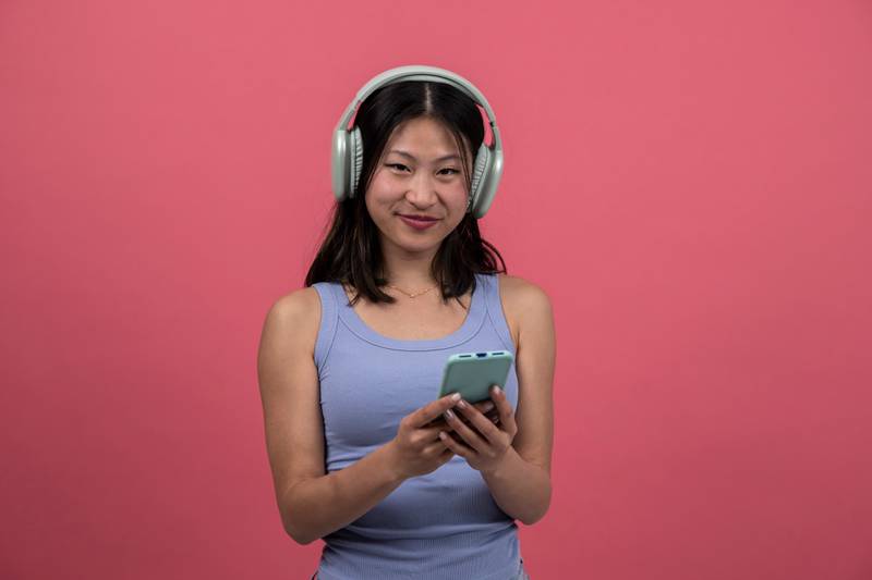 Joyful and satisfied woman with headphone texting with phone looking to the camera. Smiling young female listening to music and using online smartphone isolated on a red background.