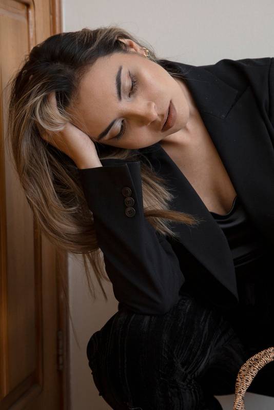 business woman with straight hair, wears makeup and a black suit, sits worried with her hand on her head,