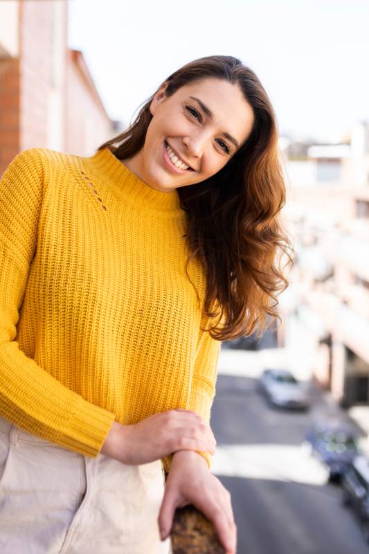 Close up of young smiling girl at leaning on a balcony on a sunny day. Cheerful woman staring at camera with a cute smile against an urban background.