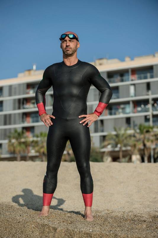 Confident man wearing a wetsuit standing in the shore ready for swimming. Professional athlete staring at the ocean in the beach with urban background.